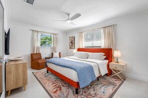 Une chambre à coucher comprenant un lit, une chaise, une table de chevet, une lampe, un ventilateur de plafond et une fenêtre avec des rideaux.