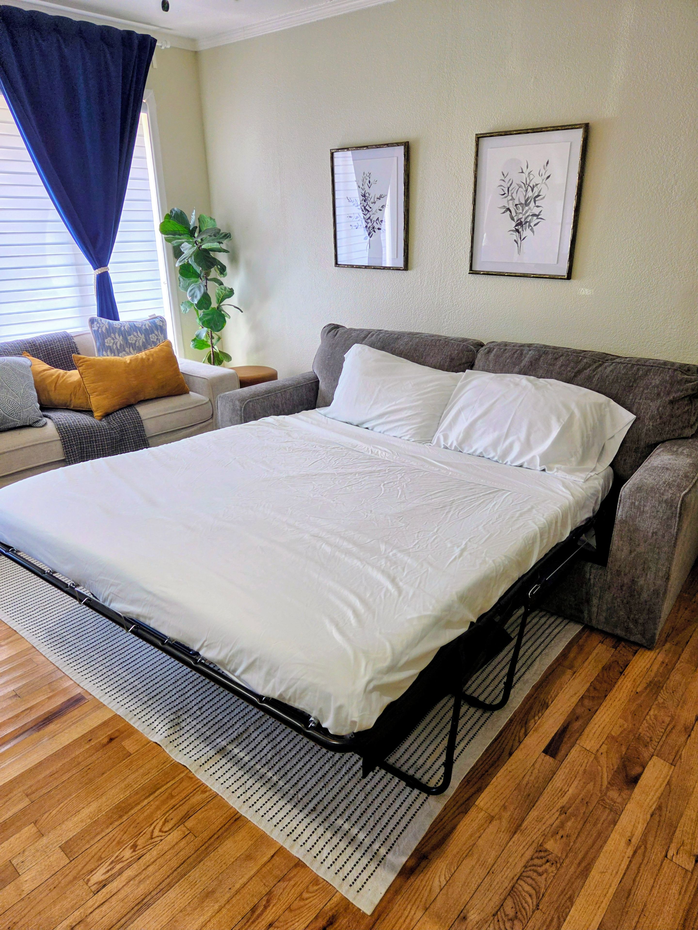 A neatly made bed with a white comforter and pillows, a grey sofa with cushions, a small side table with a plant, and two framed pictures on the wall.