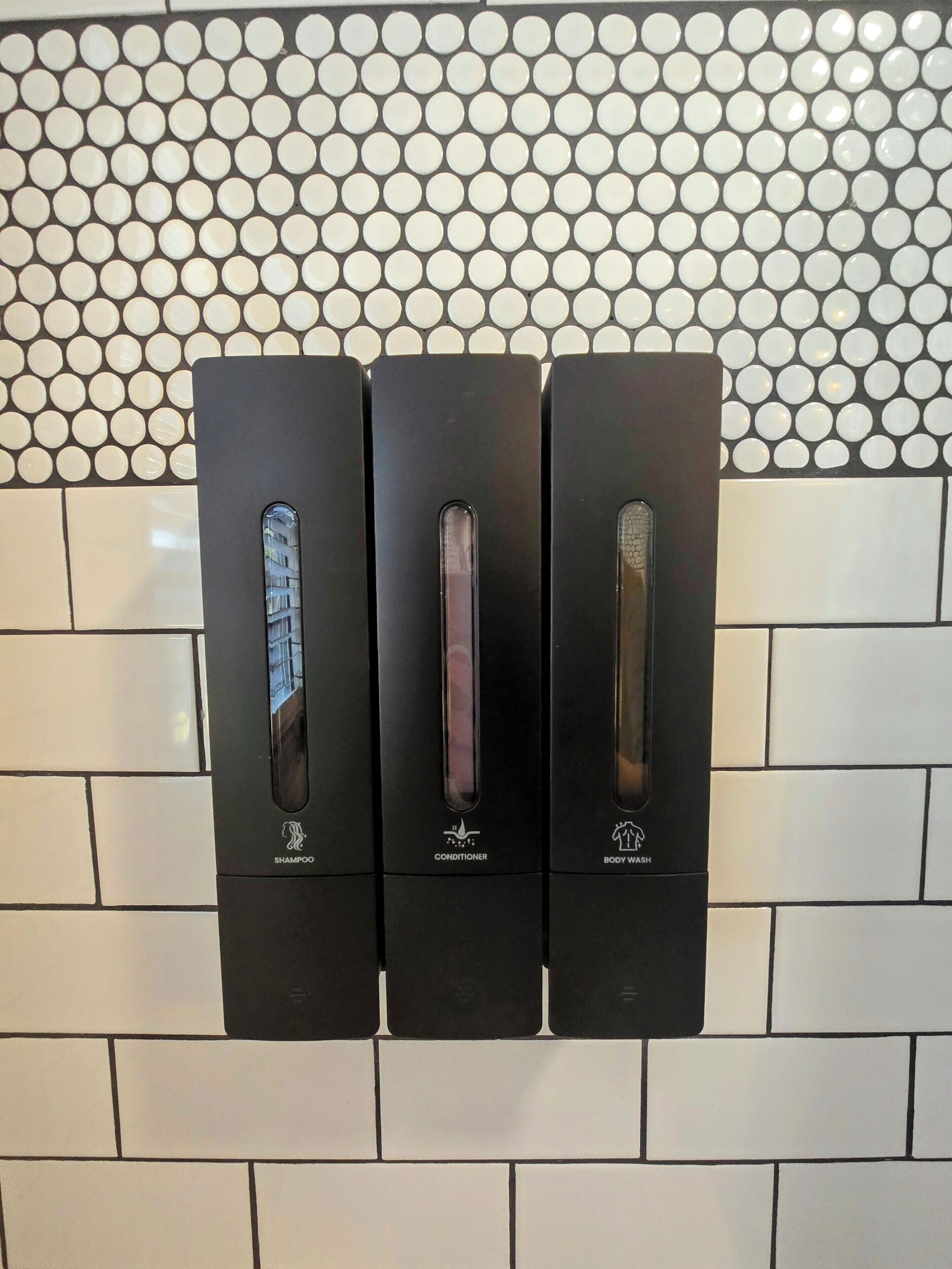 Dispensador montado en la pared con tres compartimentos etiquetados para champú, acondicionador y gel de ducha.