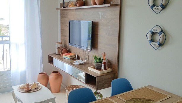 A dining area with a wooden table, blue chairs, and a TV mounted on a wooden wall. There are decorative items on the wall and a window with curtains. - Apartamento Refugio das Ondas - Piscina (Palhoça)