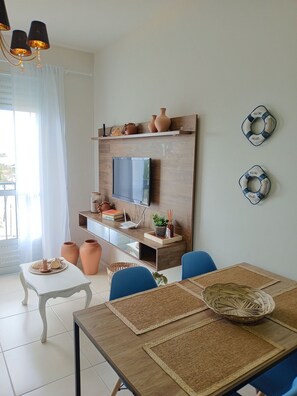 A dining area with a wooden table, blue chairs, and a TV mounted on a wooden wall. There are decorative items on the wall and a window with curtains. - Apartamento Refugio das Ondas - Piscina (Palhoça)