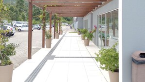 A covered walkway with potted plants, a tiled floor, and a building with large windows. - Apartamento Refugio das Ondas - Piscina (Palhoça)