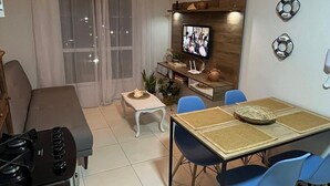 A modern living room with a sofa, dining table with chairs, a TV mounted on the wall, and a kitchen area with a stove. - Apartamento Refugio das Ondas - Piscina (Palhoça)