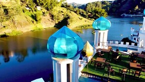 A riverside view of a hotel with two prominent blue-domed towers, a pool area, and a dining terrace. - Hotel y Termales Castillo del Lago (El Peñol)