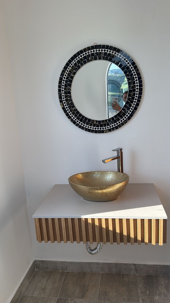 Une salle de bain avec un miroir rond, un lavabo en forme de coupe doré et un meuble-lavabo en bois.