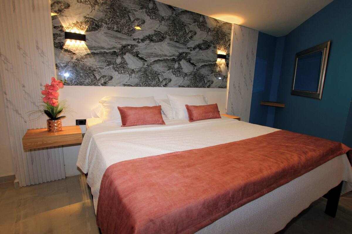 A bedroom with a large bed, a marble-patterned headboard, a nightstand with a vase of flowers, and a wall-mounted picture frame.