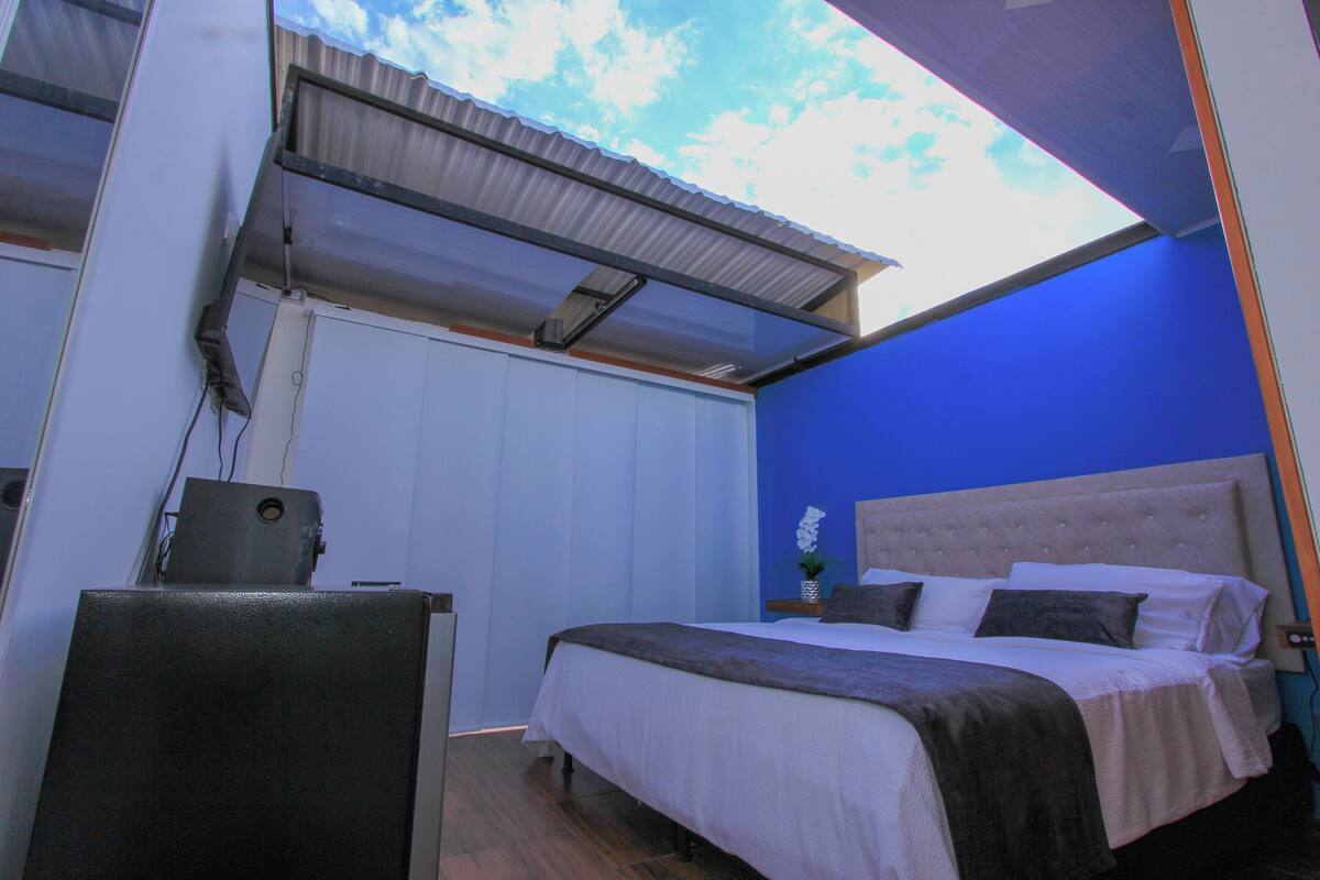 A bedroom with a bed, a desk, and a window with a view of the sky.