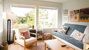 A living room with a sofa, chairs, a coffee table, and a floor lamp. There are windows with a view of trees and a wall with framed artwork.