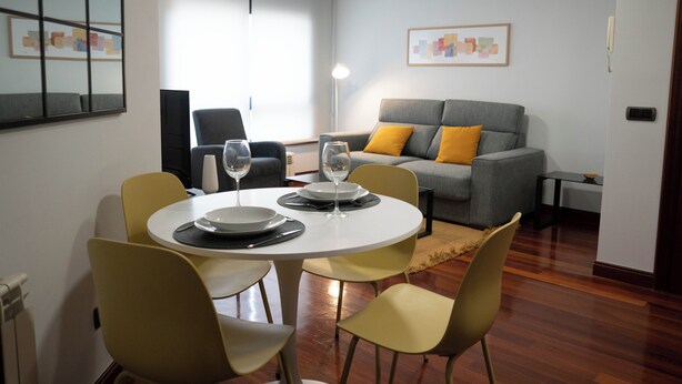 A modern living room with a dining table set for two, a grey sofa with yellow cushions, and a large window with blinds. - Centrally located apartment in Vigo 5 minutes from Puerta del Sol (Vigo)
