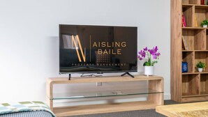 A modern living room with a flat-screen TV on a wooden stand, a glass coffee table, a sofa, and a bookshelf with books and a small plant.