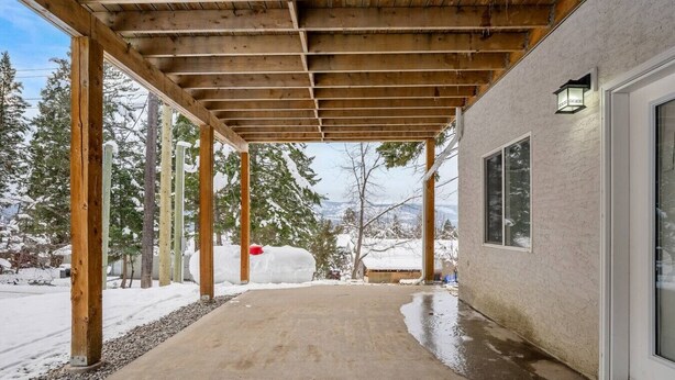 A covered outdoor area with a concrete floor, wooden beams, and a building with windows and a door.