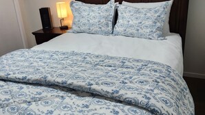 Ein ordentlich gemachtes Bett mit einer blau-weißen Bettdecke und passenden Kissen, ein Holz-Kopfstück, ein gerahmtes Bild an der Wand und ein Nachttisch mit Lampe und Telefon.