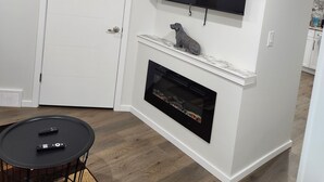 Ein modernes Wohnzimmer mit Kamin, einem Wand-Fernseher und einem kleinen Tisch mit Fernbedienungen.