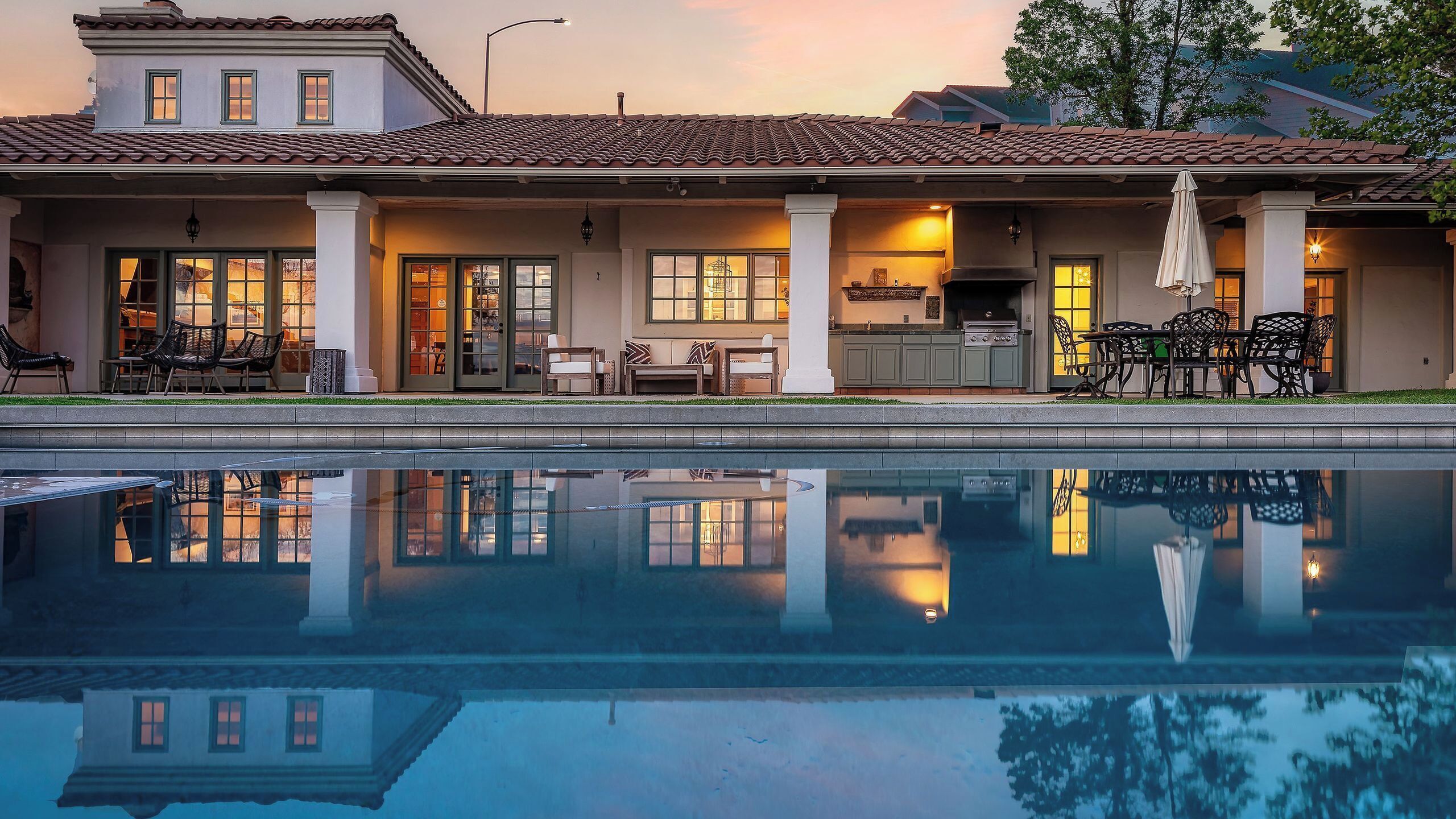 A pool with a reflection of a building, a patio with outdoor furniture, and a kitchen area.