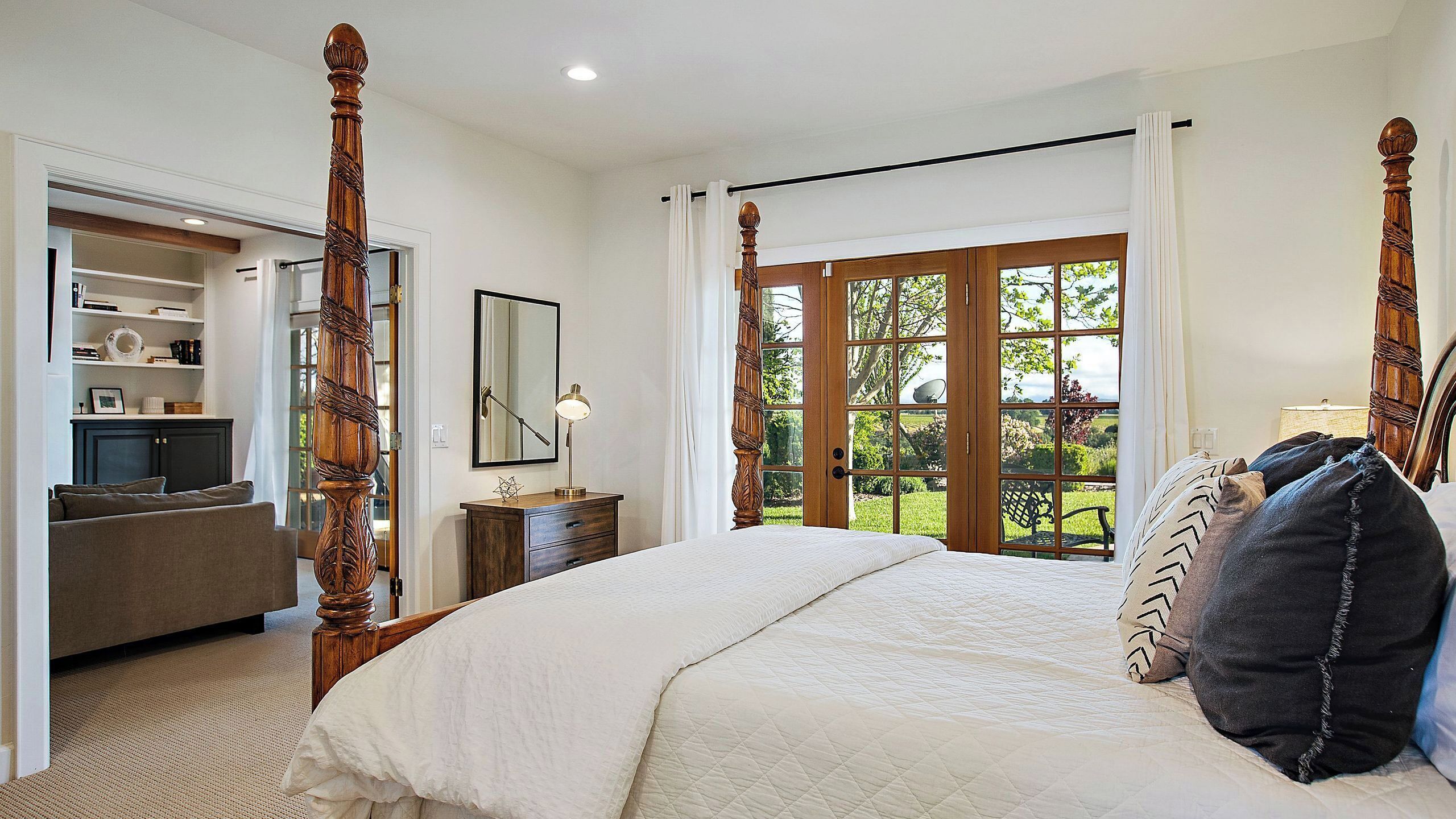 A bedroom with a four-poster bed, a desk with a lamp, a bookshelf, and a view of the outdoors.