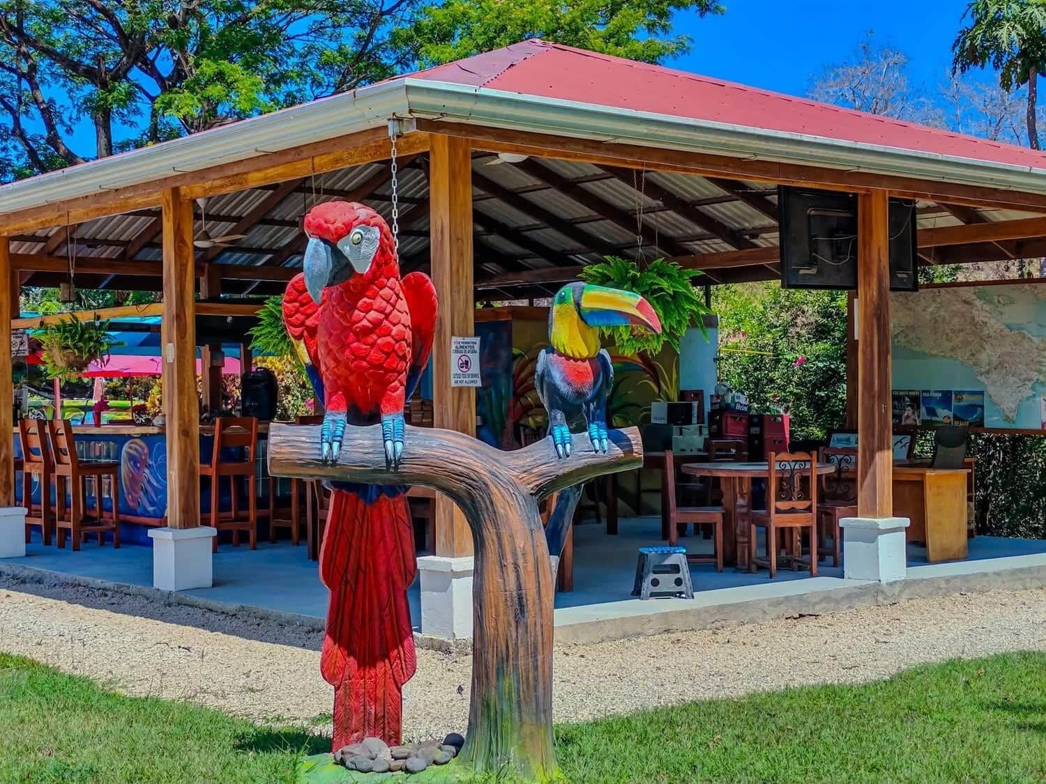 Un'area salotto esterna coperta con sedie e tavoli di legno, una struttura con tetto rosso e due grandi sculture di pappagalli su un piedistallo in legno.