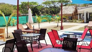 An outdoor seating area with tables and chairs, a pool, and a thatched-roof building in the background. - Tuki Tuki Lodge, Tuki Tuki Lodge - Samara (Sámara)