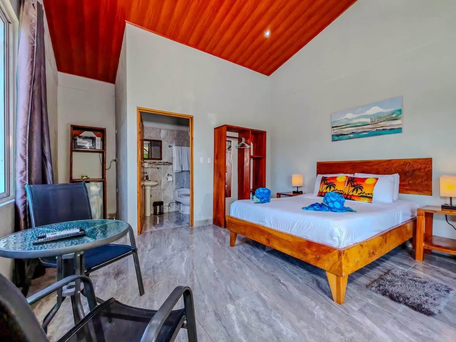 Una habitación de hotel con cama, una silla, una mesa, un baño y un cuadro en la pared.