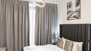 A hotel room with a bed, grey curtains, a headboard, and a painting on the wall. - Azizi Riviera Studio with Chic Furnishing (Dubai)