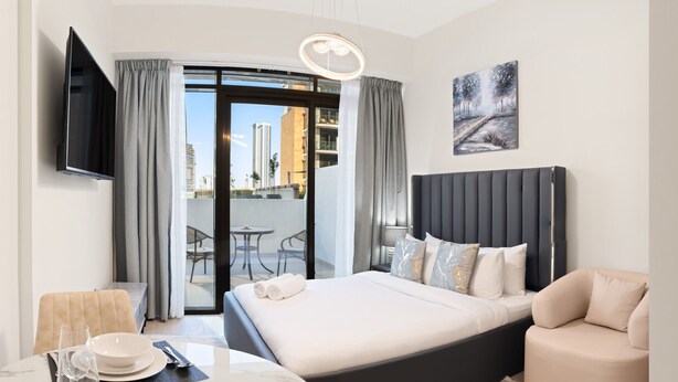 A modern hotel room with a bed, a TV, a dining area with a table and chairs, and a balcony with a view of the city. - Azizi Riviera Studio with Chic Furnishing (Dubai)