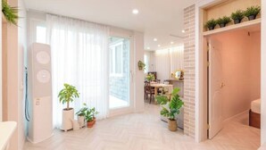 Un'ampia zona giorno con una grande finestra, un'area pranzo e una cucina visibile attraverso una porta aperta. Sono presenti diverse piante in vaso e un altoparlante bianco nella stanza.