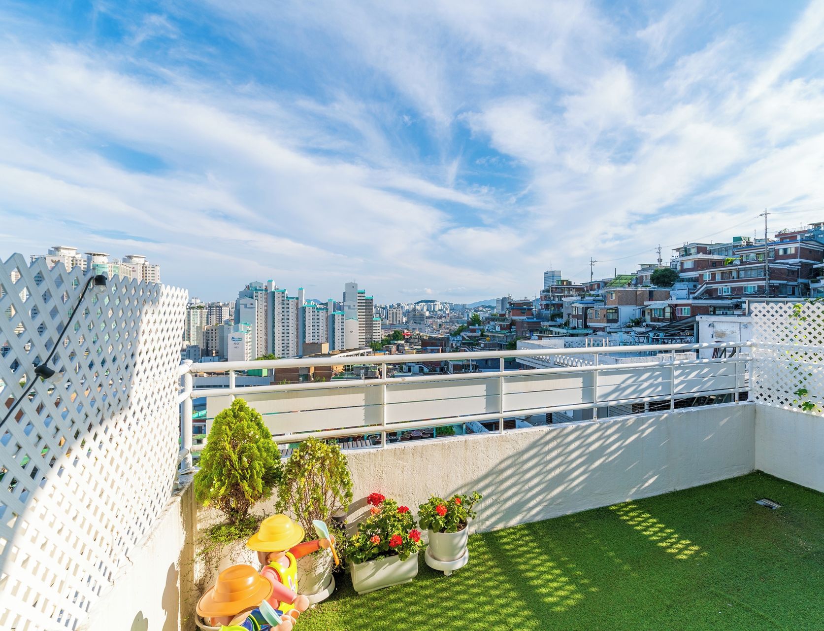 A rooftop garden with a view of a cityscape, featuring a white fence, green grass, and potted plants.
