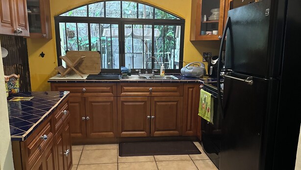 A kitchen with wooden cabinets, a black refrigerator, a window with a view of greenery, and a tiled floor. - Charming 2-bedroom townhome in welcoming Heredia with WiFi, AC in walkable town (Heredia)