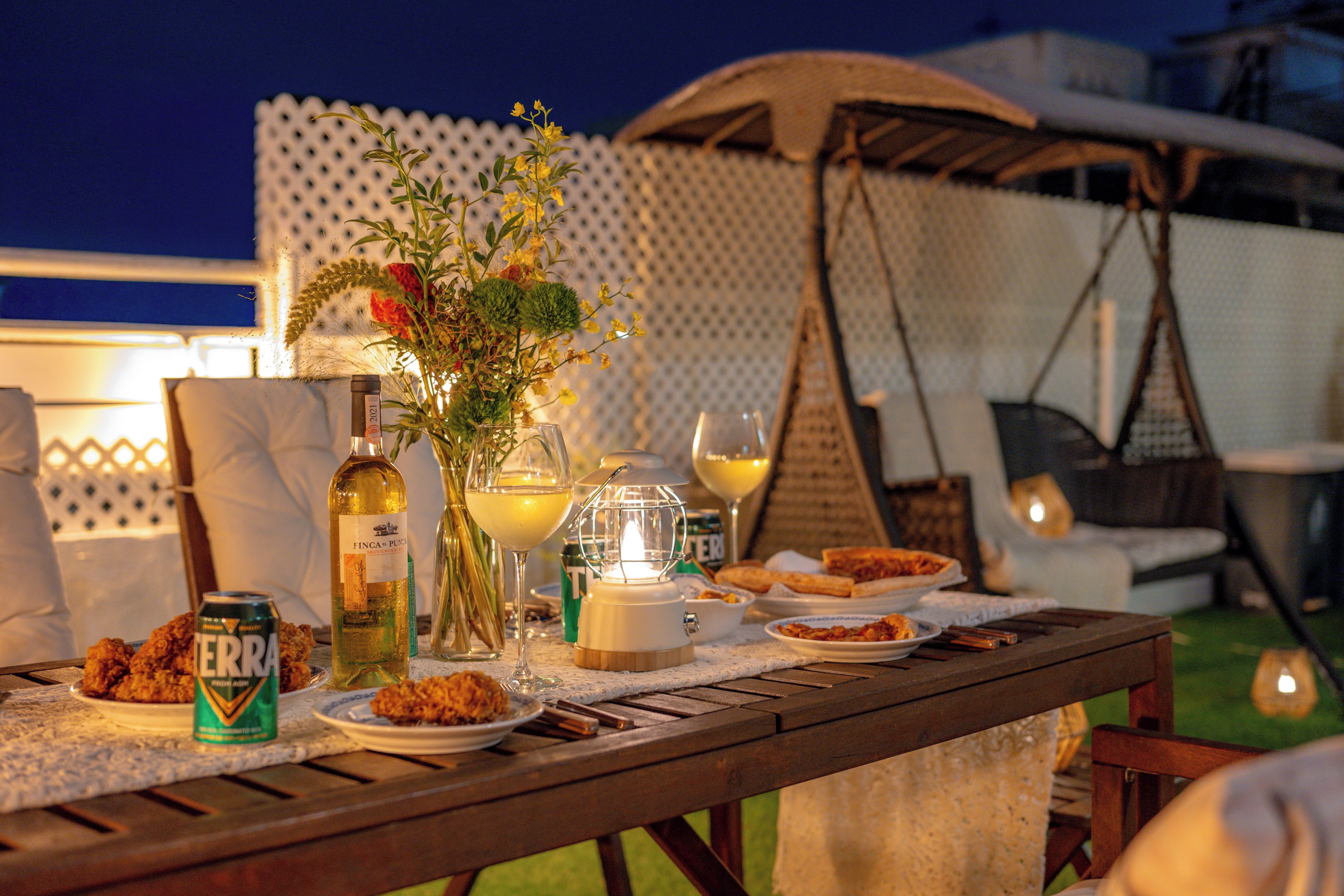 A dining table set outdoors with a bottle, glass, can, and plates of food.