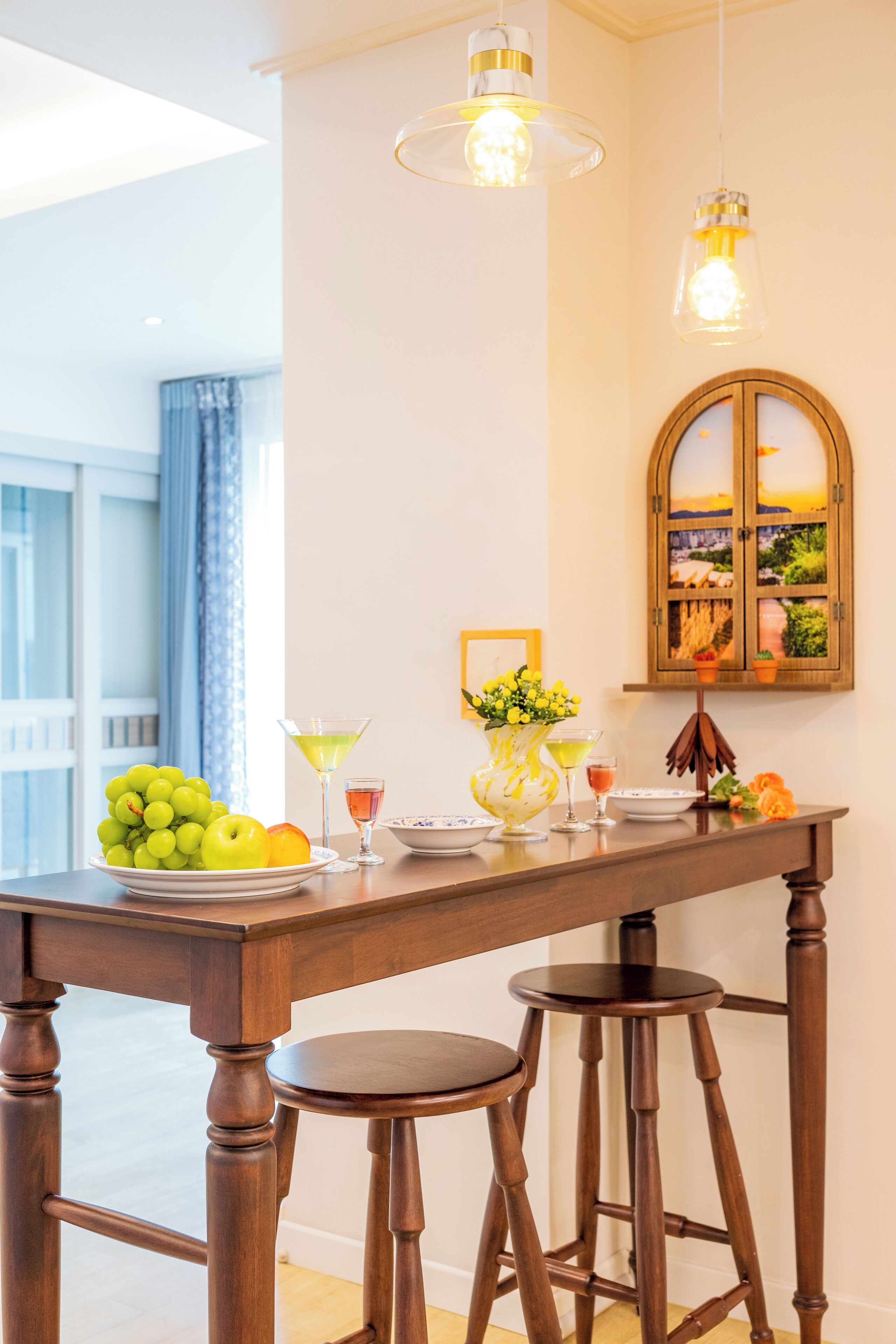 A dining area with a wooden table, two barstools, a vase with flowers, a fruit bowl, and a wall-mounted cabinet with a view.