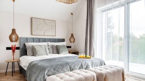 Ein modernes Schlafzimmer mit einem großen Bett, zwei Nachttischen, einer Nachttischlampe, einem Fenster mit Vorhängen und Blick ins Grüne.