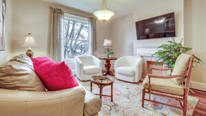 A living room with a beige sofa, two white armchairs, a wooden chair, a small round table, and a large flat-screen TV mounted on the wall.