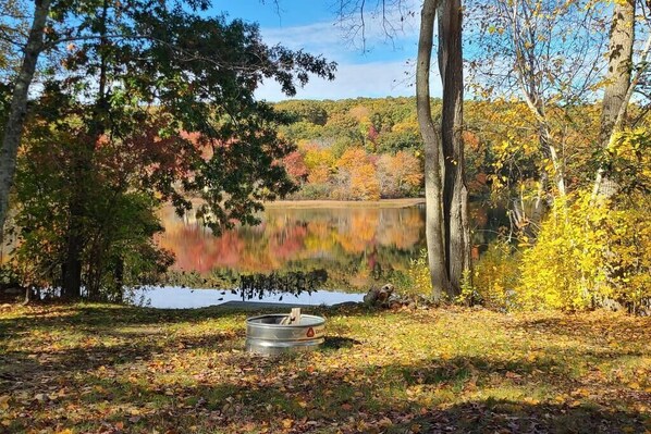 A serene lakeside setting with a campfire pit and trees reflecting on the water. - Lakefront Boho Tiny House (East Lyme)