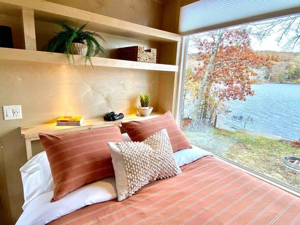 A bedroom with a large window, a bed with striped bedding, a bedside table with a plant, and a view of a lake and trees.