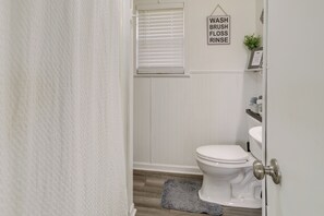 Une salle de bain comprenant des toilettes, un lavabo, une douche avec une porte blanche, et une pancarte au-dessus du lavabo indiquant : « SE LAVER, SE BROSSEZ LES DENTS, UTILISEZ DU FIL DENTAIRE, RINCEZ-VOUS LA BOUCHE ».