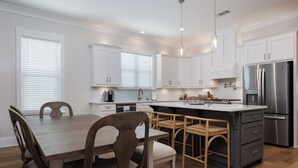 A modern kitchen with white cabinets, a stainless steel refrigerator, and a wooden dining table with chairs. - 3BR/3BA perfect location in Ocean Springs-walk to downtown-Moondance on Magnolia (Ocean Springs)