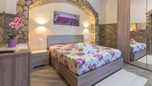 Un dormitorio con arco de piedra, una cama con ropa de cama floral, una mesita de noche, un espejo y una orquídea en maceta.