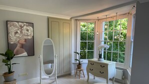 Une chambre à coucher comprenant un lit, une commode, une coiffeuse avec un miroir, une plante en pot et une fenêtre avec des rideaux.