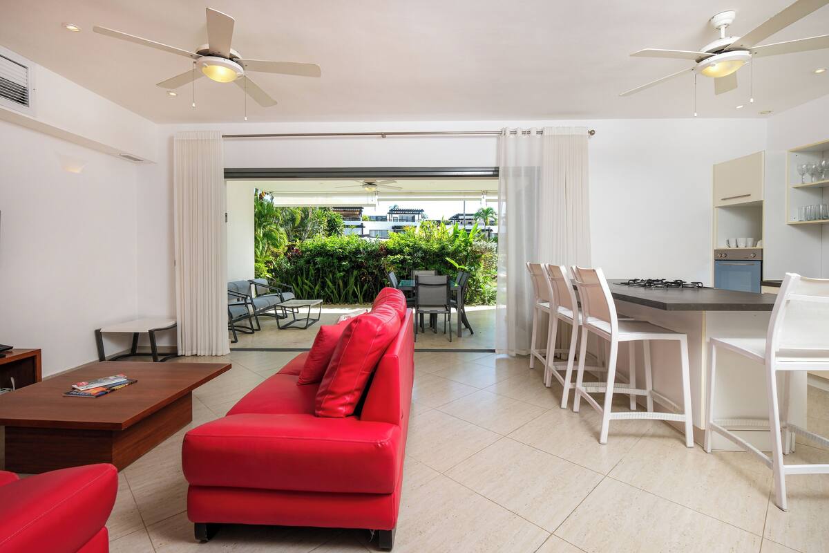A modern living room with a red sofa, a wooden coffee table, and a dining area with white chairs and a kitchen counter.