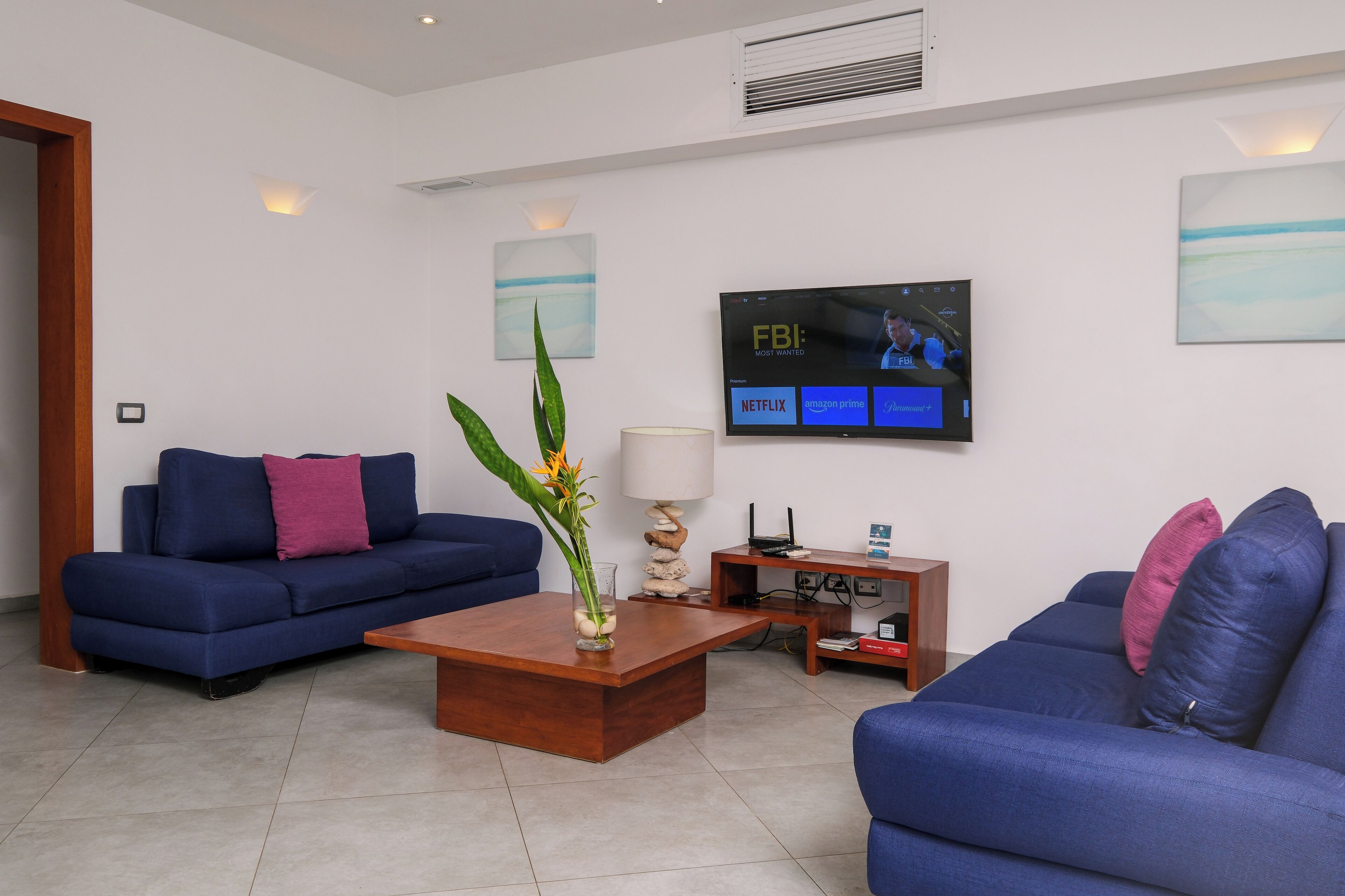 A modern living room with a blue sofa set, a wooden coffee table, and a flat-screen TV displaying a program.