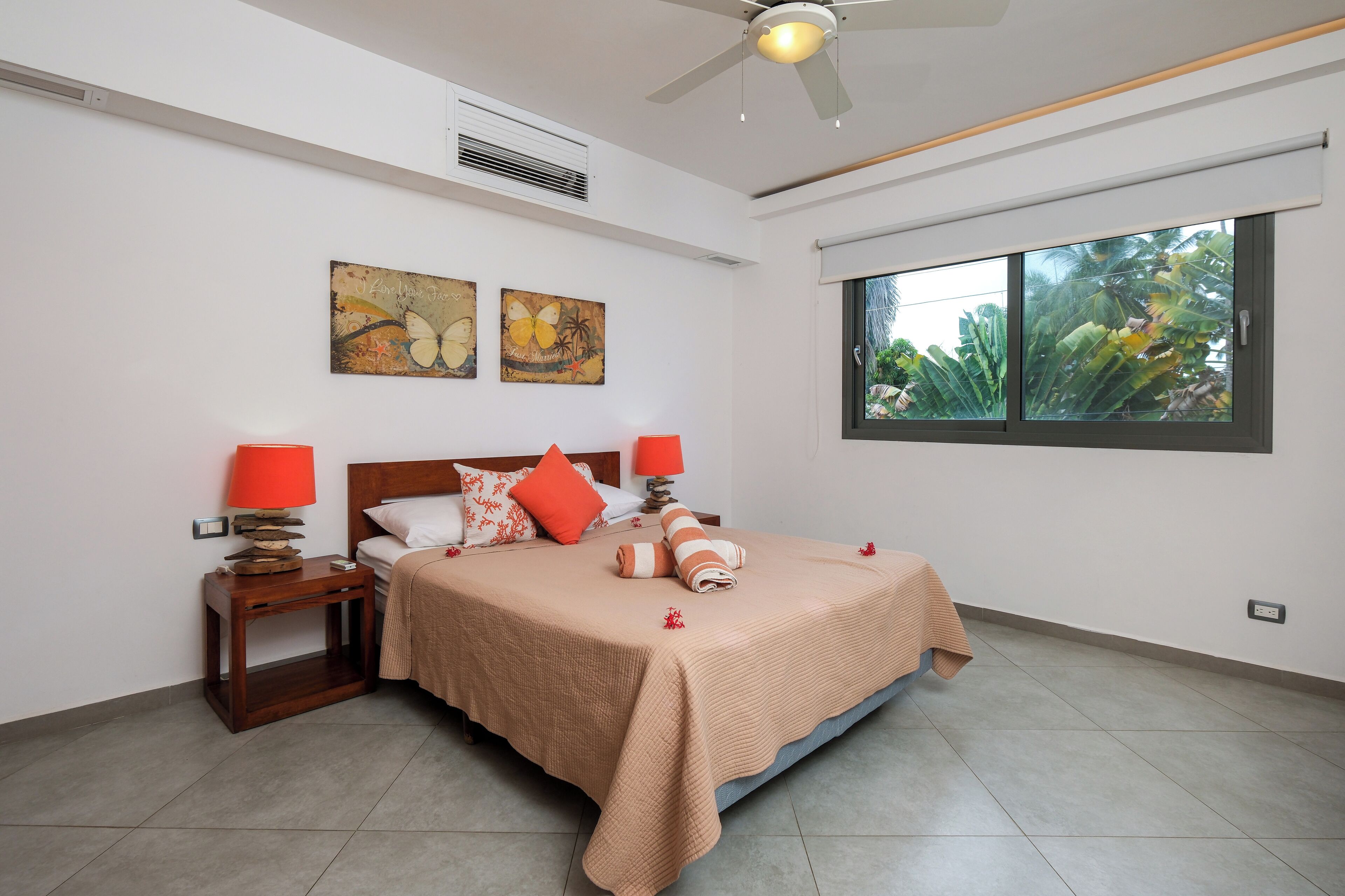 Une chambre à coucher bien aménagée, avec un lit, deux tables de chevet, un ventilateur de plafond et une grande fenêtre donnant sur un paysage verdoyant.
