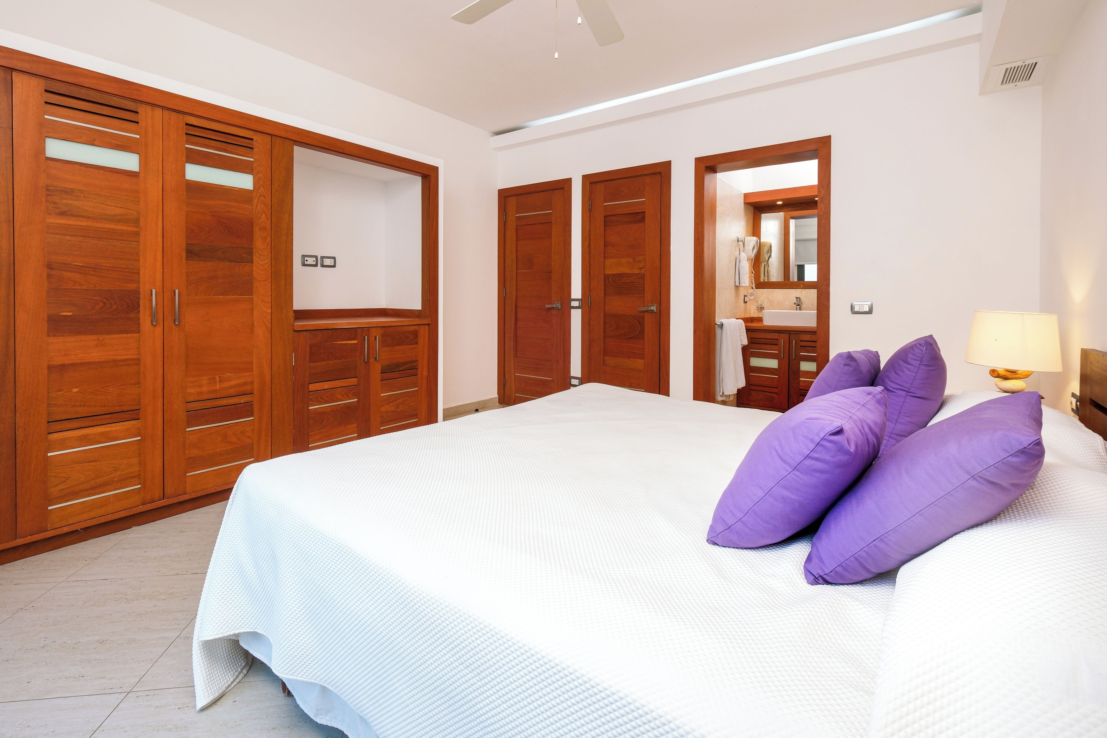 A bedroom with a large bed, wooden wardrobes, a bathroom with a mirror, and a ceiling fan.