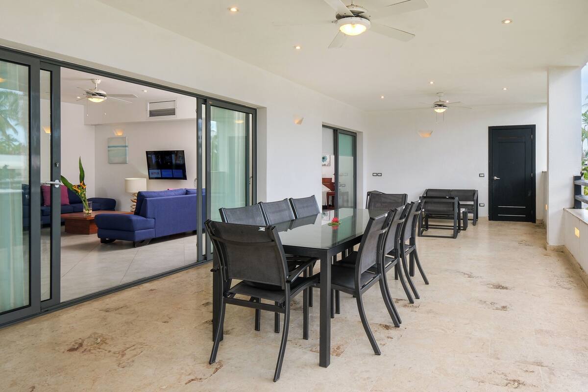 A modern dining area with a glass table and chairs, a ceiling fan, and a TV mounted on the wall.