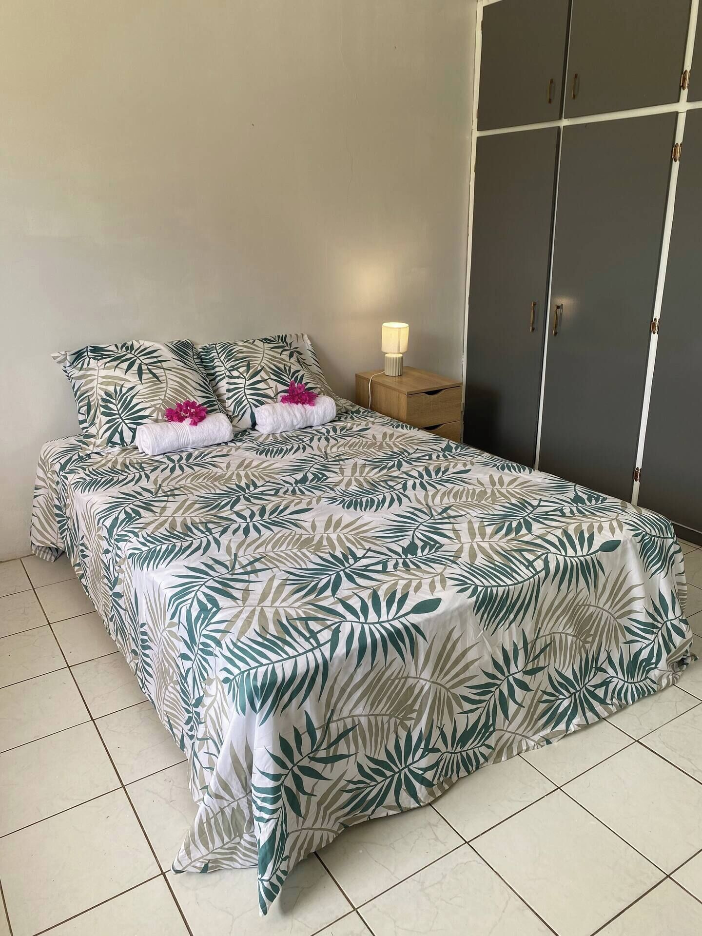 Un lit bien fait, recouvert d’une couvre-lit à motifs tropicaux, une table de chevet en bois avec une lampe et une armoire grise.