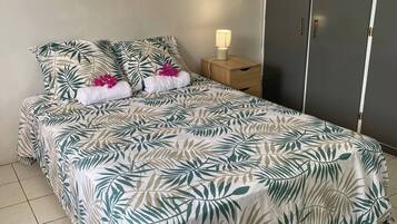Un lit bien fait, recouvert d’une couvre-lit à motifs tropicaux, une table de chevet en bois avec une lampe et une armoire grise.