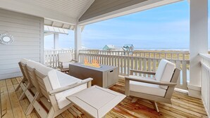 A covered outdoor deck with white furniture, a fire pit, and a view of the beach.