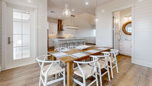 A spacious dining area with a wooden table and white chairs, a kitchen in the background, and a large mirror on the wall.