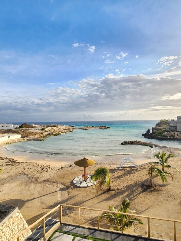 A beach with a clear view of the sea, a sandy area with a palm tree, and a thatched umbrella.