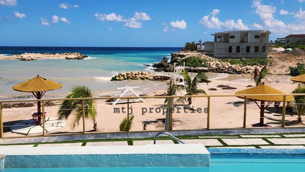 Vista a la piscina de un resort frente a la playa, con un cielo azul despejado, arena y un edificio al fondo.