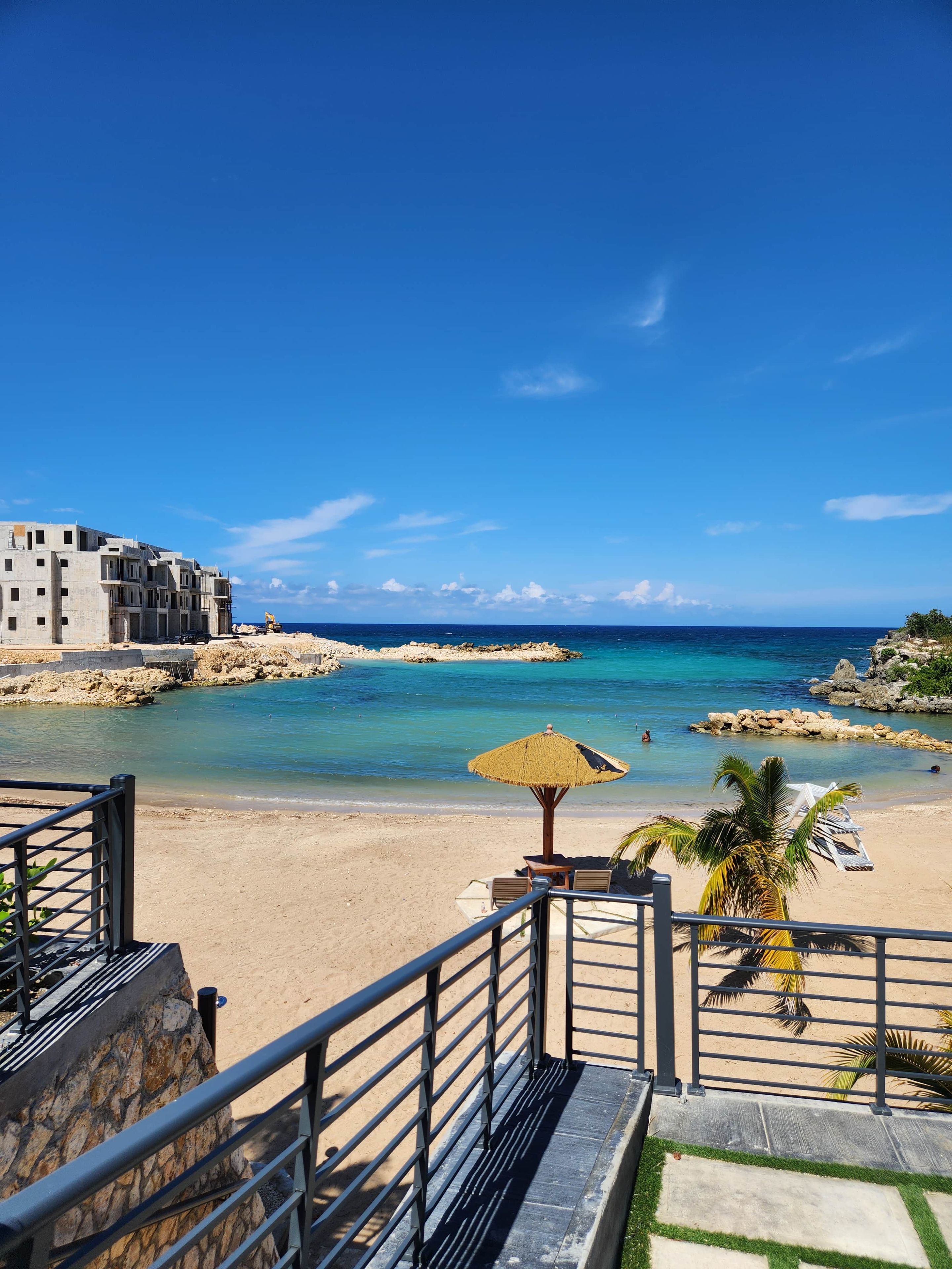 A beach view with a clear sky, a sandy shore, and a building with a balcony overlooking the sea.