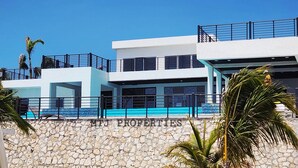 Un edificio moderno de varios pisos con fachada de piedra, un balcón con barandilla negra y un cielo azul despejado.
